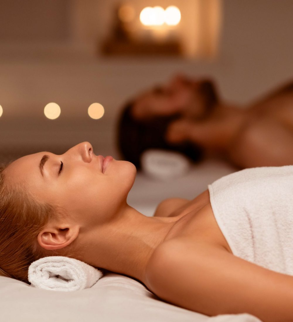 Couple Resting After Massage In Luxury Spa With Candles, Relaxing Lying With Eyes Closed, Wrapped In Towels During Aromatherapy In Beauty Salon Indoor. Romantic Spa Day And Relaxation. Selective Focus